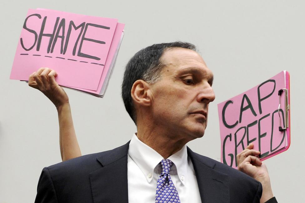 File photo of protestors holding signs behind Fuld as he takes his seat to testify at a House Oversight and Government Reform Committee
